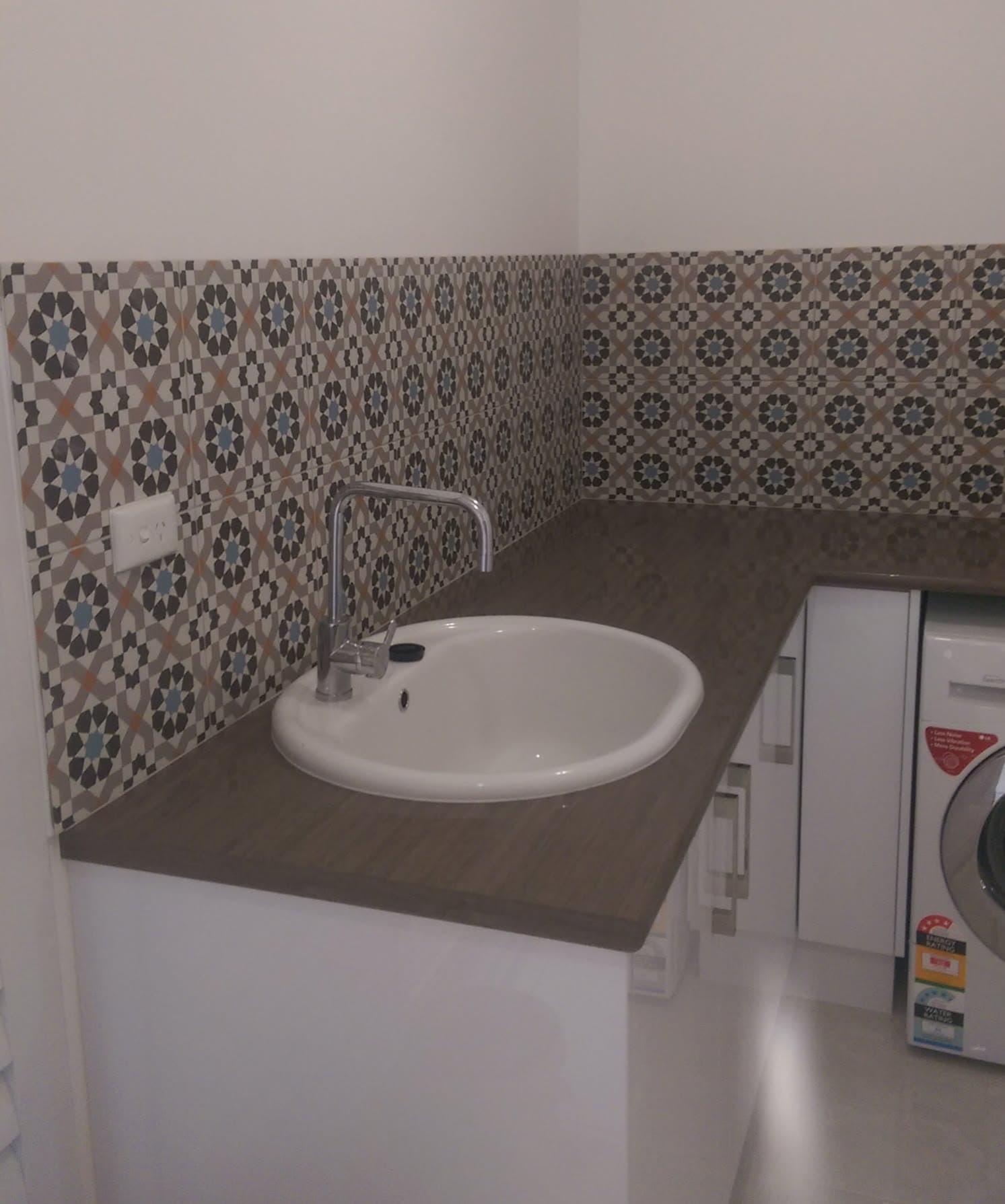 Grey, black and blue patterned tile laundry splashback above a trough sink, with a washing machine alongside